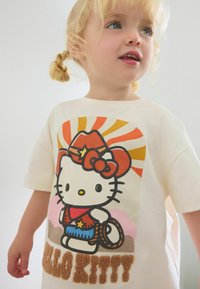 Kremowa koszulka z nadrukiem charakteru z kreskówki w czerwonym kapeluszu kowbojskim z kokardką, z dużym nadrukiem graficznym oraz puszystym napisem "HELLO KITTY" na dole.