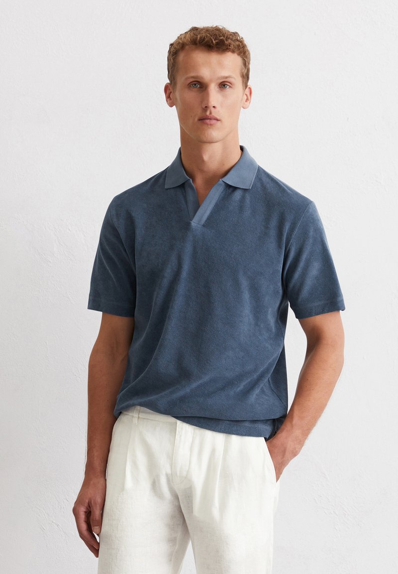 Polo shirt in donkerblauwe stof met korte mouwen, een v-hals ontwerp en een contrasterende lichtblauwe kraag. G gedragen met lichtgekleurde broeken.