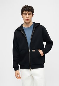 Νέος άνδρας με σγουρά σκούρα μαλλιά, φορώντας μαύρο φούτερ με φερμουάρ πάνω από μπλε μπλούζα και λευκό παντελόνι, με το ένα χέρι στην τσέπη.