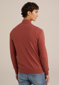 Pulover tricotat de culoare portocalie arsă, cu un guler înalt, mâneci lungi și manșete cu nervuri. Purtați peste blugi de un albastru deschis, cu o textură netedă.