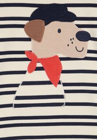 Koft med bomull som har et hundeapplikasjon med brunt øre og svart hatt, rød bandana, mot en bakgrunn med kremfargede og marineblå striper.