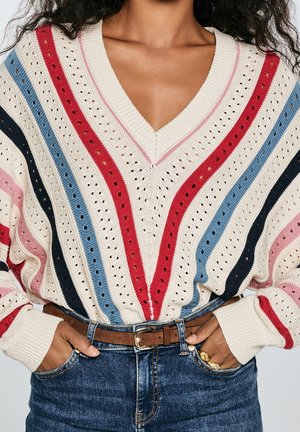 Jersey de cuello en V color crema con rayas rojas, azules, negras y rosas en un patrón de punto perforado, metido dentro de unos vaqueros azules con cinturón marrón.