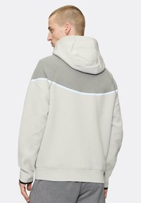 Sudadera gris claro con un panel gris más oscuro en la parte superior de la espalda, que presenta una línea de acento blanca. Tejido suave y corte relajado, con capucha.