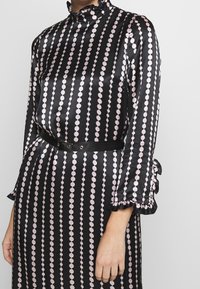 Vestido de satén negro con un patrón circular rosa, cuello alto, puños con volantes y un cinturón negro que acentúa la cintura.