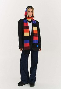 Czarna marynarka na białym topie, do tego wielokolorowy, paskowany szalik. Szerokie dżinsy o fasonie z rozszerzanymi nogawkami i czarne obuwie dopełniają wygląd.