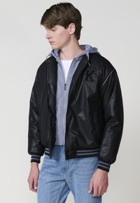 Giacca bomber nera con una texture lucida, dotata di zip frontale, polsini a coste, dettagli a strisce e un logo sul petto. Indossata sopra un felpa grigia.