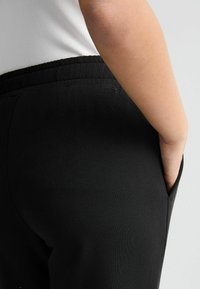 Pantalons de survêtement noirs en tissu doux, dotés d'une taille élastique et d'une poche latérale. Le design est simple, sans motifs ni accents visibles.