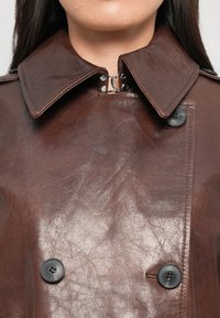 Gros plan sur une personne portant un trench-coat en cuir marron foncé avec un large col, des boutons noirs à double boutonnage et un fermoir en métal au niveau du cou.