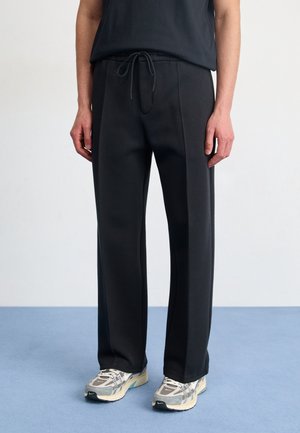 Person trägt schwarze, gerade geschnittene Hosen mit Kordelzugbund und graue-weiße Sportschuhe, steht auf blauem Boden vor weißer Wand.