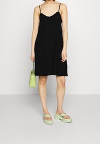 Černé šaty na tenkých ramínkách se zvlněným výstřihem, doplněné zelenými sandály na podpatku a odpovídající zelenou kabelkou. Hladká textilie.