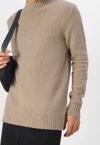 Mann trägt einen beigen strukturierten Strickpullover und schwarze Hosen, hält den schwarzen Schultertaschenriemen über die linke Schulter.