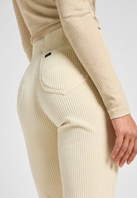 Pantaloni in corda a coste color crema con vita alta, dotati di tasche posteriori e dettagli di cucitura. Mano appoggiata sul lato della gamba.