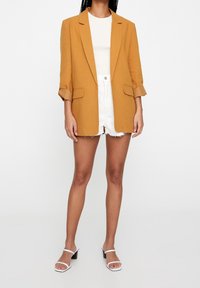 Blazer oversize jaune moutarde avec une texture lisse, manches retroussées et deux poches avant, associé à un short blanc et des sandales.