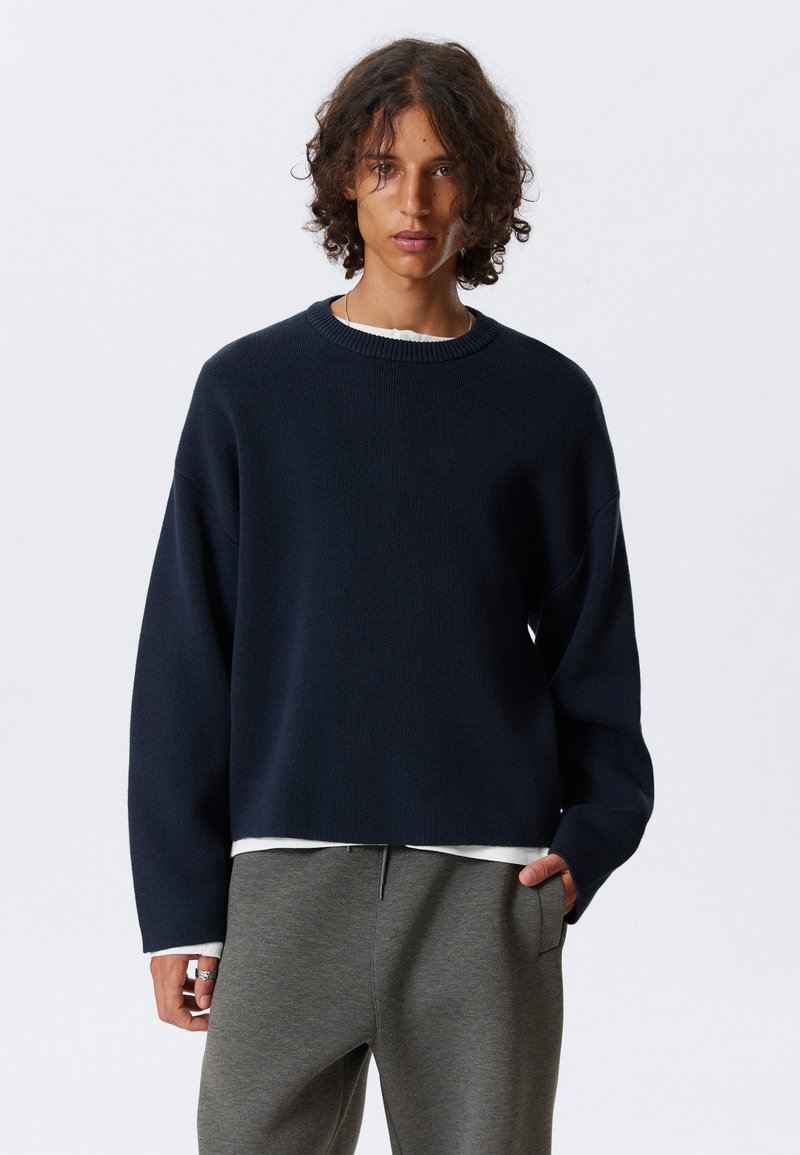 Ung person med lockigt hår har en marinblå tröja över en vit skjorta och grå sweatpants, stående mot en enfärgad ljus bakgrund.