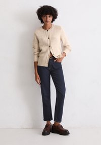 Beige gebreide cardigan met een ronde halslijn en knoopsluiting, gecombineerd met donkere blauwe rechte jeans en bruine veterschoenen.