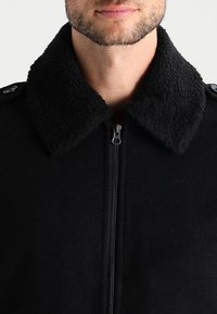 Svart zip-up jacka med en texturerad svart fårskinnskrage, med knapparamarkeringar och en tät passform. Mjuk, slät tygtextur.