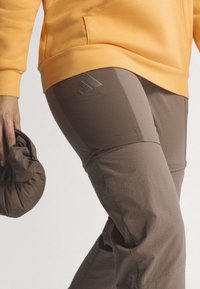 Personne portant un pantalon marron avec le logo Adidas et un sweat jaune, tenant un accessoire en tissu marron froissé dans une main.