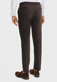 Brązowe spersonalizowane spodnie o gładkiej fakturze, z dwiema tylnymi kieszeniami i eleganckimi liniami, w połączeniu z ciemnobrązowymi zamszowymi butami.