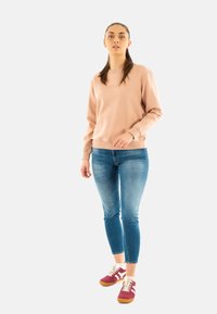 Felpa rosa chiaro con collo rotondo e polsini a costine, abbinata a jeans blu aderenti e scarpe da ginnastica borgogna con dettagli bianchi.