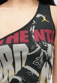 Débardeur noir côtelé avec un décolleté diagonal, présentant un graphisme imprimé d'un joueur de basketball, des lettres rouges et grises, et un accent de logo jaune.