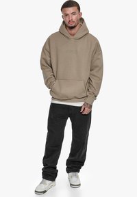 Beige oversized hoodie med kengurulomme, parret med løse sorte bukser og hvide sneakers. Enkel design med diskret branding.