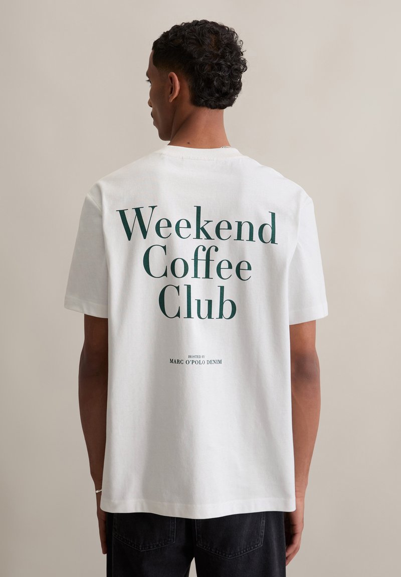 Witte katoenen T-shirt met korte mouwen, met grote groene tekst op de achterkant met de tekst "Weekend Coffee Club," en kleinere tekst eronder.