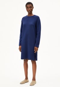 Robe tricotée à manches longues couleur marine avec un col rond et des fentes latérales, assortie à des chaussures beiges à enfiler. Texture plate, coupe au niveau du genou.