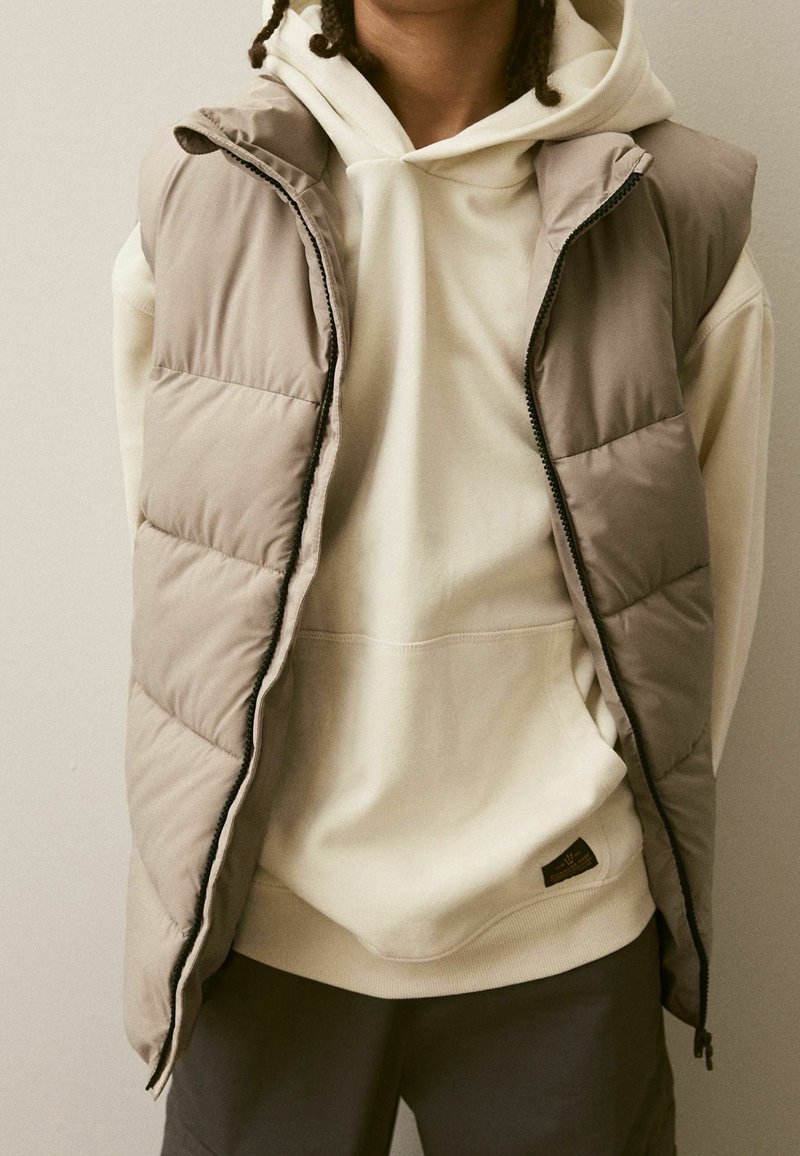 Personne portant un sweat à capuche crème et un gilet matelassé beige avec un pantalon foncé, debout devant un fond clair uni.