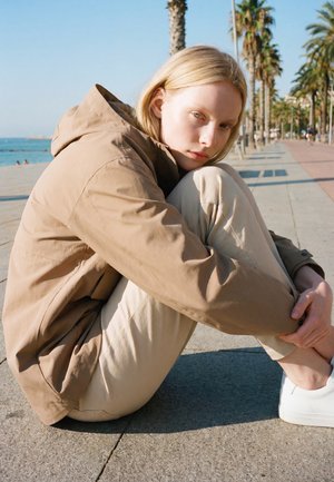 Une femme blonde vêtue d’une veste et d’un pantalon beige est assise, les genoux serrés contre elle, sur une promenade en bord de mer ensoleillée bordée de palmiers.