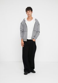 Pelēka rāvējslēdzēja kapuces krekls ar baltu oderi, apvienots ar vienkāršu baltu T-kreklu un brīvām melnām biksēm. Modelis stāv pretī baltai fona.