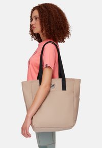 Sac fourre-tout beige avec des poignées noires, texture lisse et logo discret. Comprend plusieurs poches et une forme rectangulaire structurée.