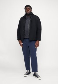 Black jacket with a high collar, grey knit sweater underneath, and navy trousers; model wears black and white high-top sneakers.
