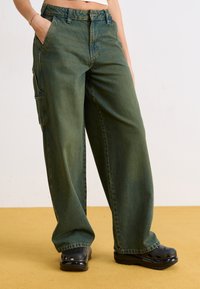 Pantaloni cargo verde deschis cu o croială lejeră, din material denim, cu buzunare laterale și un design cu picior drept. Asortați cu saboți negri.