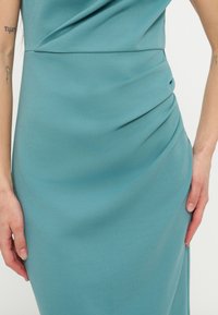 Γυναίκα που φοράει στενό φόρεμα σε τιρκουάζ απόχρωση με λεπτομέρειες ραμμένες στη μέση, εμφανίζοντας τατουάζ στα αντιβράχια, με τα χέρια να ακουμπούν στα πλάγια.