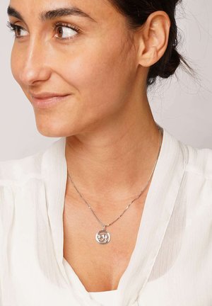 Woman with dark hair tied back wearing a white blouse and a silver necklace with a round pendant showing a figure inside.