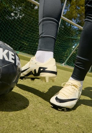 Piłkarz w białych korkach Nike, gotowy do kopnięcia czarno-białej piłki na zielonym boisku z siatką bramki.