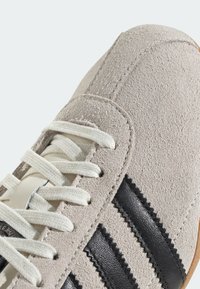 Beige suède sneakers med vita snörningar, med tre svarta läderränder och en texturerad yta. Texturerade detaljer nära sulan.