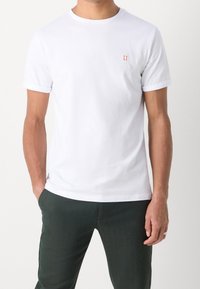 Mann trägt ein schlichtes weißes T-Shirt mit kurzen Ärmeln und einem kleinen roten "II"-Logo auf der Brust, kombiniert mit dunkelgrünen Hosen, die Hände in den Taschen.