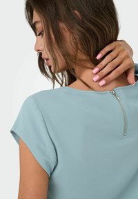 Frau mit braunen Haaren und pinken N�ägeln, die ein hellblaues Oberteil mit Reißverschluss auf dem Rücken trägt, berührt ihren Hals und schaut nach links vor einem weißen Hintergrund.