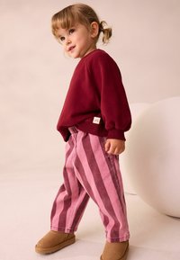 Rød sweatshirt, stribede bukser i pink og burgundy, samt tan støvler. Blødt stof med løs pasform og ribbede manchetter. Mærket synligt på sweatshirten.