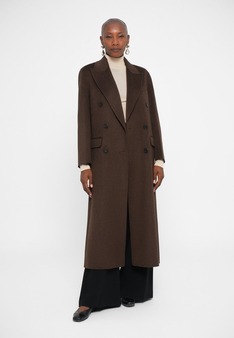 Manteau en laine marron à double boutonnage avec de grands boutons noirs, présentant une silhouette longue et droite avec des revers en pointe, porté par-dessus un haut beige.