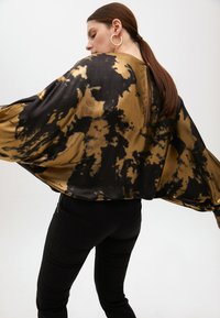 Blusa nera e oro in stile tie-dye con maniche ampie e dettaglio della cerniera sul retro, abbinata a pantaloni neri aderenti. Tessuto liscio e lucido.