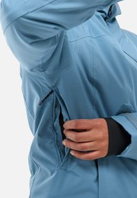 Light blue waterproof jacket with a zippered side pocket, featuring a smooth texture, adjustable cuffs, and a streamlined design.