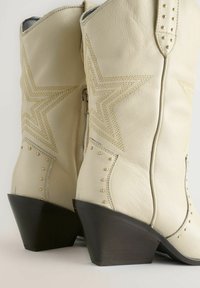 Vita lädercowboystövlar med stjärnmönster, gyllene sömmar, nitar och en mörk trä, vinklad klack. Dragkedjeslutning bak.