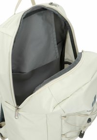 Mochila de color beige claro con forro interior gris, gran apertura con cremallera, bolsillo frontal y correas ajustables. Presenta un logo sutil y un compartimento de almacenamiento.