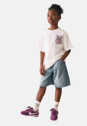 Young girl standing with hand in pocket, wearing oversized white graphic t-shirt, denim shorts, white socks, and purple Nike sneakers.