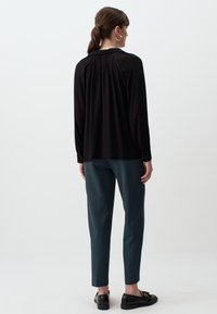 Haut noir à manches longues avec un dos plissé, associé à un pantalon tapered vert foncé et des chaussures noires, sur un fond clair.