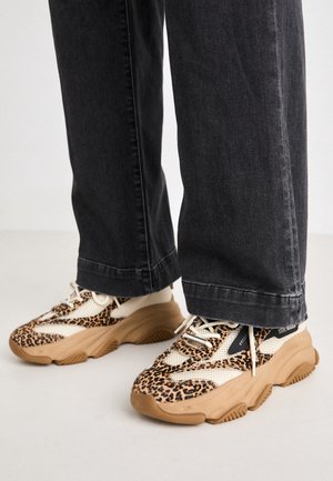 Baskets épaisses à imprimé léopard et détails en maille beige portées avec un jean large noir sur un sol blanc.