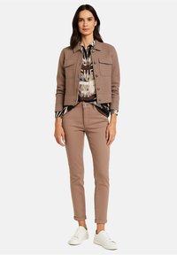 Jasnobrązowa kurtka krótka z przednimi kieszeniami, noszona na wzorzystą bluzkę, w połączeniu z jasnobrązowymi dopasowanymi dżinsami i białymi sneakersami.