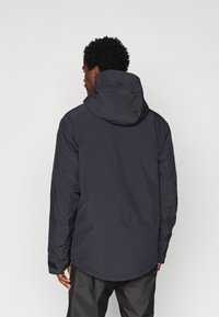 Homem com cabelo curto e encaracolado, vestindo uma jaqueta impermeável preta com capuz e calças pretas, visto de costas contra um fundo simples.